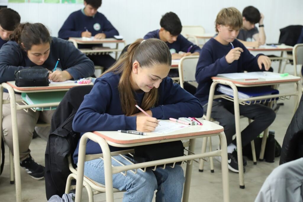 Alumnas y alumnos de secundaria con uniforme azul resolviendo una evaluación en sus bancos, en un aula luminosa.