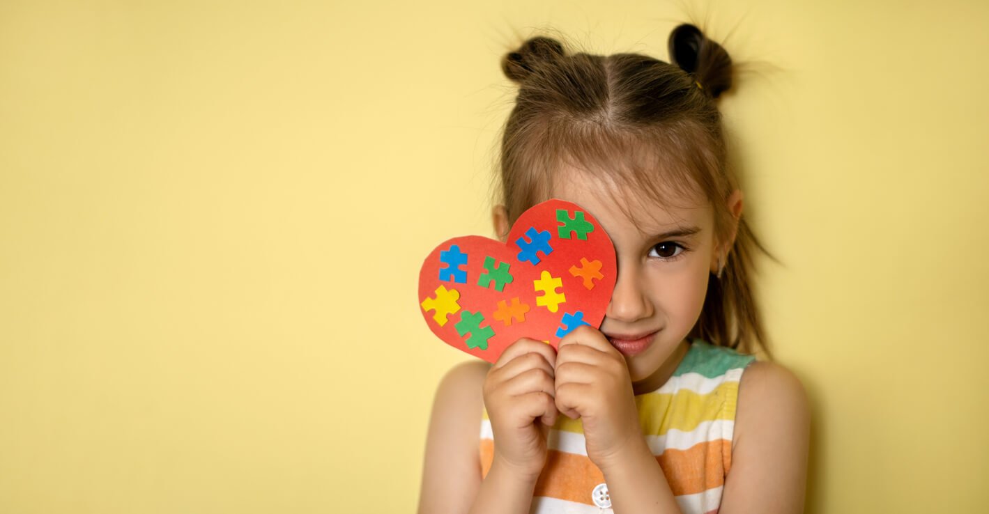 Niña sonriente que se cubre un ojo con un corazón de papel rojo decorado con piezas de rompecabezas de colores, símbolo de la concientización sobre el autismo.